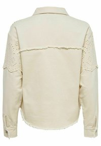Veste chemise beige avec col, ornements en dentelle sur les épaules, ourlet effiloché et poignets à boutons. Le tissu semble texturé et léger.