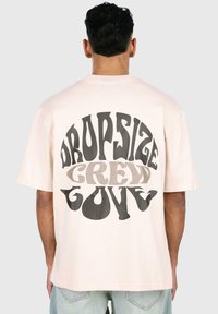 Hellrosa übergroßes T-Shirt aus Baumwolle. Der Rücken zeigt ein grafisches Textdesign in dunkelgrau und beige mit der Aufschrift "DROPSIZE CREW GOVE."