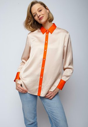 Satijnen blouse in lichtbeige met een oranje kraag en knoopsluiting. Lange mouwen met oranje manchetten en een ontspannen pasvorm.