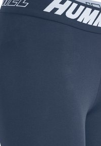Leggings blu navy con una ampia vita elastica che presenta la scritta "HUMMEL" in bianco. Tessuto liscio e elasticizzato con un design aderente.