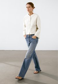 Kvinde, der går indendørs iført en hvid tekstureret jakke med knapper, blå straight-leg jeans og sorte sandaler med åbne tæer og firkantede hæle.