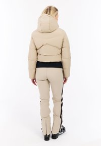 Kvinde iført en beige pufferjakke med hætte, beige bukser med sorte side-striber og sorte skistøvler, som vender bort fra en hvid baggrund.