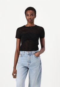 Svart kortermet topp laget av et teksturert, gjennomsiktig stoff med horisontale striper. Kombinert med lyseblå høyhalsede denimjeans.