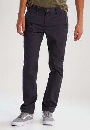 Mężczyzna ubrany w ciemnogranatowe spodnie o prostych nogawkach, szaro-białe trampki, stojący z rękami opuszczonymi wzdłuż ciała na jednolitym tle.