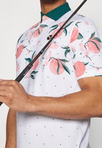 Bílý polo tričko s růžovými květinovými vzory a zelenými akcenty. Obsahuje malé zelené tečky a černé logo značky. Nosič má golfovou hůl.