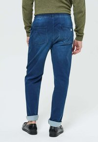 Pantalon en denim bleu avec une coupe décontractée, taille élastique, deux poches arrière et ourlets retroussés, accompagné de chaussures noires sur un fond uni.