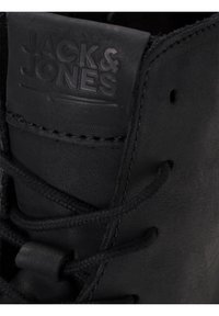 Close-up van een zwart leren schoen, met het reliëflogo "JACK & JONES" en veters die door de gaatjes zijn geregen.