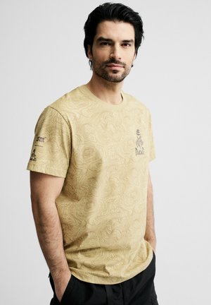 Mężczyzna o ciemnych włosach i brodzie, ubrany w beżowy wzorzysty t-shirt z logo Dakar, stoi z rękami w kieszeniach na jednolitym tle.