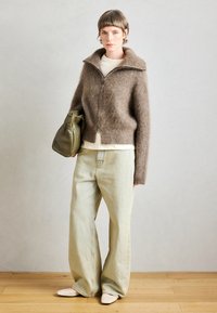 Cardigã castanho felpudo com fecho, camisa creme, calças largas verde claro e sapatos brancos sem atacadores. Bolsa verde-oliva na mão.