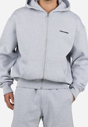 Mann trägt hellgrauen Kapuzenpullover mit Reißverschluss und passende Jogginghose, mit "PEGADOR"-Logo auf der linken Brust des Hoodies.