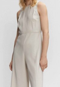 Combinaison sans manches beige avec un design droit, texture lisse et fermeture éclair latérale invisible ; coupe ajustée avec un léger éclat.