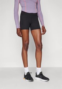 Persona vestita con pantaloni sportivi neri, maglia a maniche lunghe viola con cuciture arancioni, calze bianche e scarpe da corsa nere, in piedi contro uno sfondo bianco.