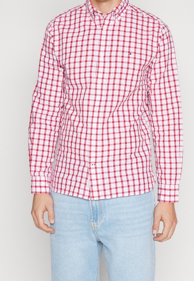 Homme portant une chemise à manches longues à carreaux rouges et blancs boutonnée, assortie à un jean bleu clair, sur un fond uni.