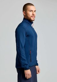 Mann in blauer, strukturierter Zip-Jacke mit hohem Kragen und seitlichem Reißverschluss, der seitlich vor einem schlichten grauen Hintergrund steht.