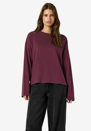 Giovane donna con lunghi capelli castani che indossa una camicia bordeaux larga a maniche lunghe e pantaloni neri a gamba larga, in piedi su sfondo bianco.