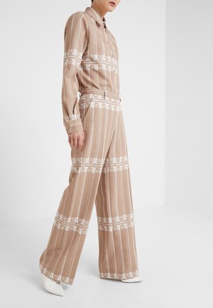 Person trägt beige und weiß gemustertes Langarmhemd und weit geschnittene Hose, kombiniert mit weißen spitzen Schuhen, steht vor einem schlichten weißen Hintergrund.