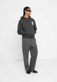 Μαύρη μπλούζα με κουκούλα με γραφικά μπροστά, συνδυασμένη με ελαιοπράσινα cargo παντελόνια με πλαϊνές τσέπες. Το μοντέλο φορά μαύρα γυαλιά ηλίου και αθλητικά παπούτσια.