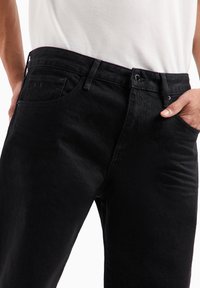 Jeans in denim nero con una texture liscia, caratterizzati da una vestibilità slim, cinque tasche tradizionali e un logo discreto sulla tasca posteriore.