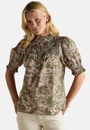 Blonde Frau, die eine grün-rosa blumige Bluse mit Puffärmeln und gesmoktem Einsatz sowie weiße Hosen trägt und mit Händen in den Taschen steht.