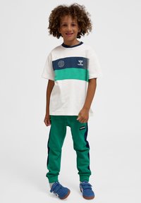 Göndör hajú gyermek fehér és türkiz Hummel pólóban, zöld melegítőnadrágban és kék sportcipőben, egyszínű háttér előtt állva.