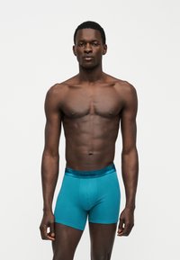 Άνδρας μοντέλο στέκεται, φοράει τιρκουάζ Calvin Klein μποξεράκια, σε απλό λευκό φόντο, με ουδέτερη έκφραση προσώπου.