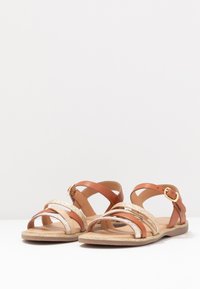 Sandalias de cuero marrón y dorado con múltiples correas, una correa con hebilla en el tobillo, acentos texturizados dorados y una suela plana color marrón claro.