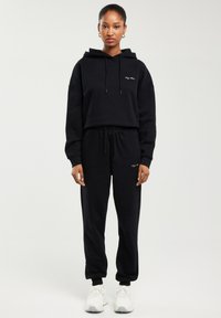 Žena stojící čelem, oblečená v černé mikině s kapucí a černých teplácích, doplněných bílými teniskami, na jednobarevném světlém pozadí.