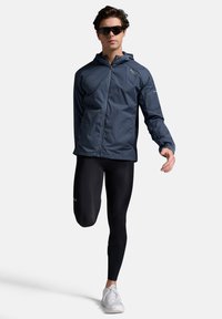 Marine windbreaker jas met een capuchon, ritsluiting; gecombineerd met zwarte leggings en witte sportschoenen. Model draagt een zonnebril.