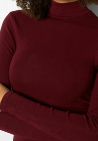 Osoba ubrana w dopasowany sweter w burgundowym kolorze z długimi rękawami, z rękami skrzyżowanymi na piersiach.