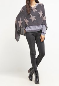 Poncho gris avec de grands motifs d'étoile beige, en matière douce, superposé à un haut bleu clair et associé à un jean skinny noir et des bottines.