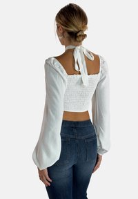 White, long-sleeve crop top with smocked back, featuring puffed sleeves and a tie detail at the neck, paired with dark blue fitted jeans.