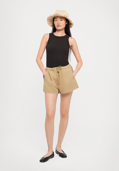 Black ribbed tank top, beige high-waisted shorts with a drawstring, and black flat shoes. Model wearing a straw hat. Neutral background.