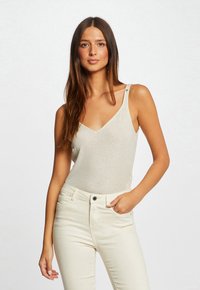 Body beige clair sans manches avec un col en V et un léger éclat ; présente deux boutons décoratifs sur l'épaule. Assorti avec un pantalon blanc.