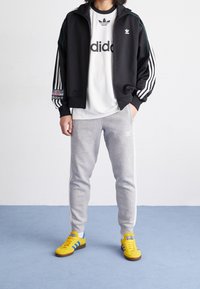 Svart zip-up jacka med vita ränder, vit t-shirt med logotyp, grå sweatpants och gula sportskor med blå accenter.