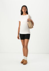 Top de maternitate alb cu dungi, pantaloni scurți negri și pantofi din suede maron deschis. Modelul poartă o geantă de culoare maron deschis. Șosetele sunt albe cu margini denivelate.