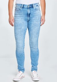 Naine, kes kannab helehalli kitsaid teksaseid ja valgeid tossusid, seisab käsivarred lõdvalt piki keha lihtsalt taustal.