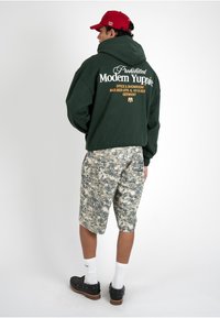 Homme portant une veste à capuche verte avec le texte « Modern Yuppie », casquette rouge, short camouflage, chaussettes blanches et chaussures noires, debout sur un fond blanc.
