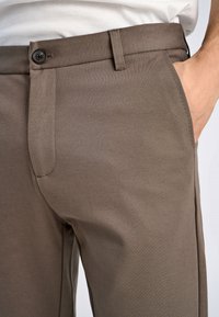Bruine stoffen broek met een gladde uitstraling, voorzien van een voorkeerzak, riemlussen en een enkele knoopsluiting bij de taille.