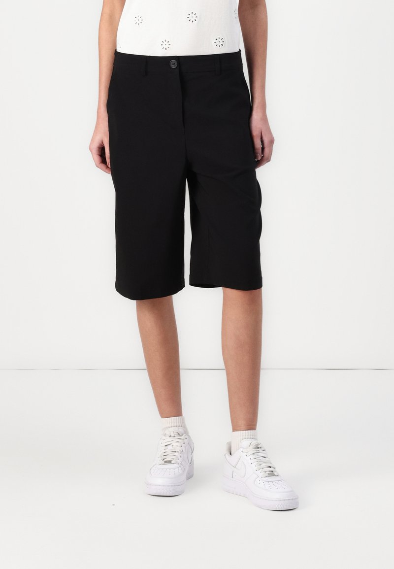 Shorts noirs jusqu'au genou en tissu lisse, avec une taille plate dotée d'une fermeture à bouton et de deux poches latérales. Portés avec des sneakers blanches.