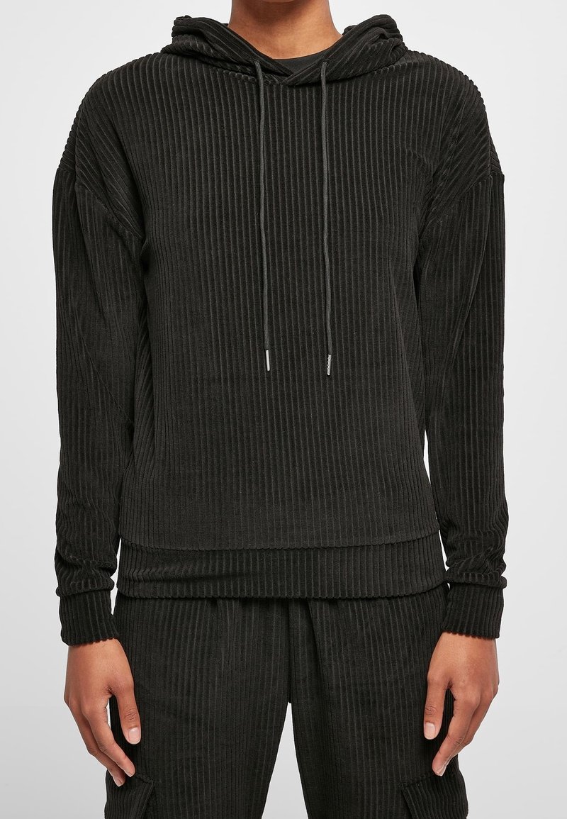 Personne portant un sweat à capuche en tissu côtelé noir et un pantalon assorti, debout les bras détendus le long du corps devant un fond uni.