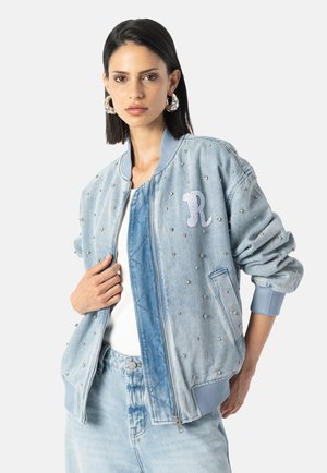 Kvinna som bär en ljusblå stentvättad jeansjacka med bokstaven "R" som patch, i kombination med matchande jeansbyxor och silverfärgade hoopsörhängen.