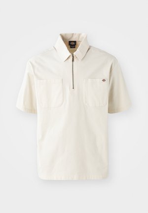 Kortærmet creme polo med lynlås krave, der har to brystlommer og et lille logo på venstre lomme. Fremstillet af en bomuldsblanding.