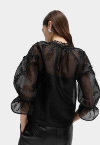 Czarna przezroczysta bluzka z kwiatową haftem, bufkami na rękawach, marszczonym dekoltem i zapięciem na guziki z tyłu. Noszona na czarnych skórzanych spodniach.