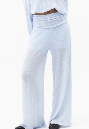 Femme portant un pantalon large texturé bleu clair et un pull court assorti, debout devant un fond blanc.