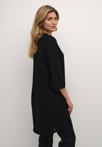 Tunique noire à manches trois-quarts, en tissu doux, coupe décontractée, fentes latérales et accents de boutons sur les manches. Design minimaliste.