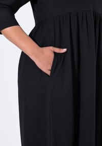 Musta mekko, joka on valmistettu pehmeästä kankaasta, jossa on tiukka yläosa ja levenevä hame. Sisältää sivutaskun ja kolmenneljäsosapituista hihaa.