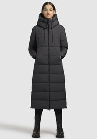 Donna in piedi che indossa un lungo cappotto invernale imbottito nero con colletto alto e cappuccio, leggings neri e stivali neri su uno sfondo semplice.