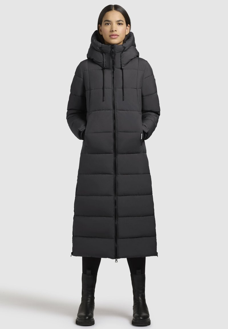 Donna in piedi che indossa un lungo cappotto invernale imbottito nero con colletto alto e cappuccio, leggings neri e stivali neri su uno sfondo semplice.