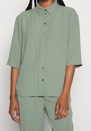 Osoba ubrana w lekko zielonawą koszulę na guziki z krótkimi rękawami o fakturze oraz dopasowane spodnie, stojąca na jednolitym tle.