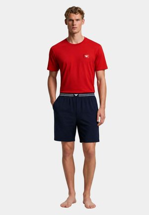 Uomo in piedi scalzo che indossa una maglietta rossa a maniche corte e pantaloncini blu navy con dettagli del logo sul petto e in vita.